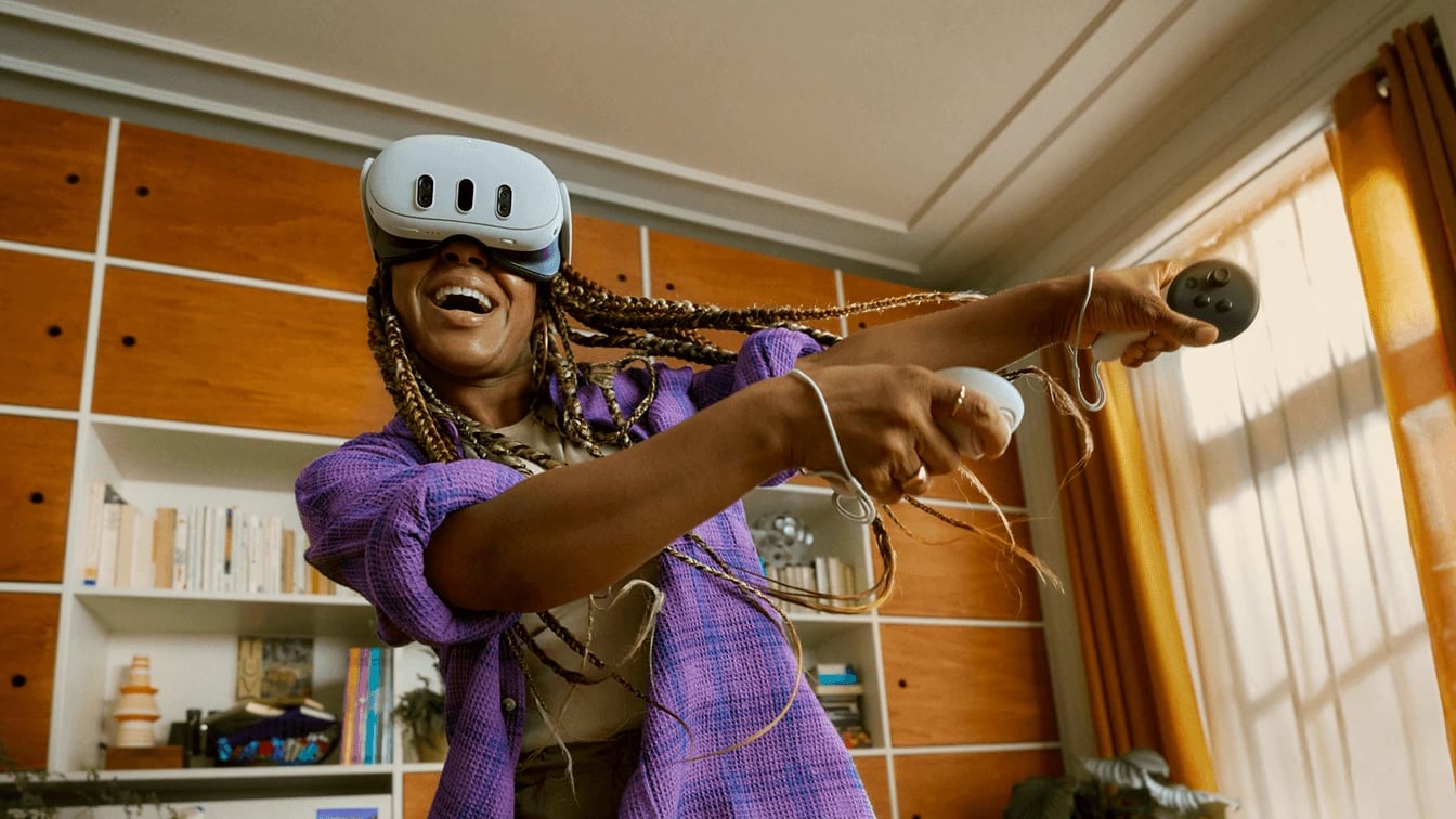 A woman laughing mid-swing in a Meta Quest 3 in her living room, both arms out with the controllers, bookshelves and an armchair well clear behind her