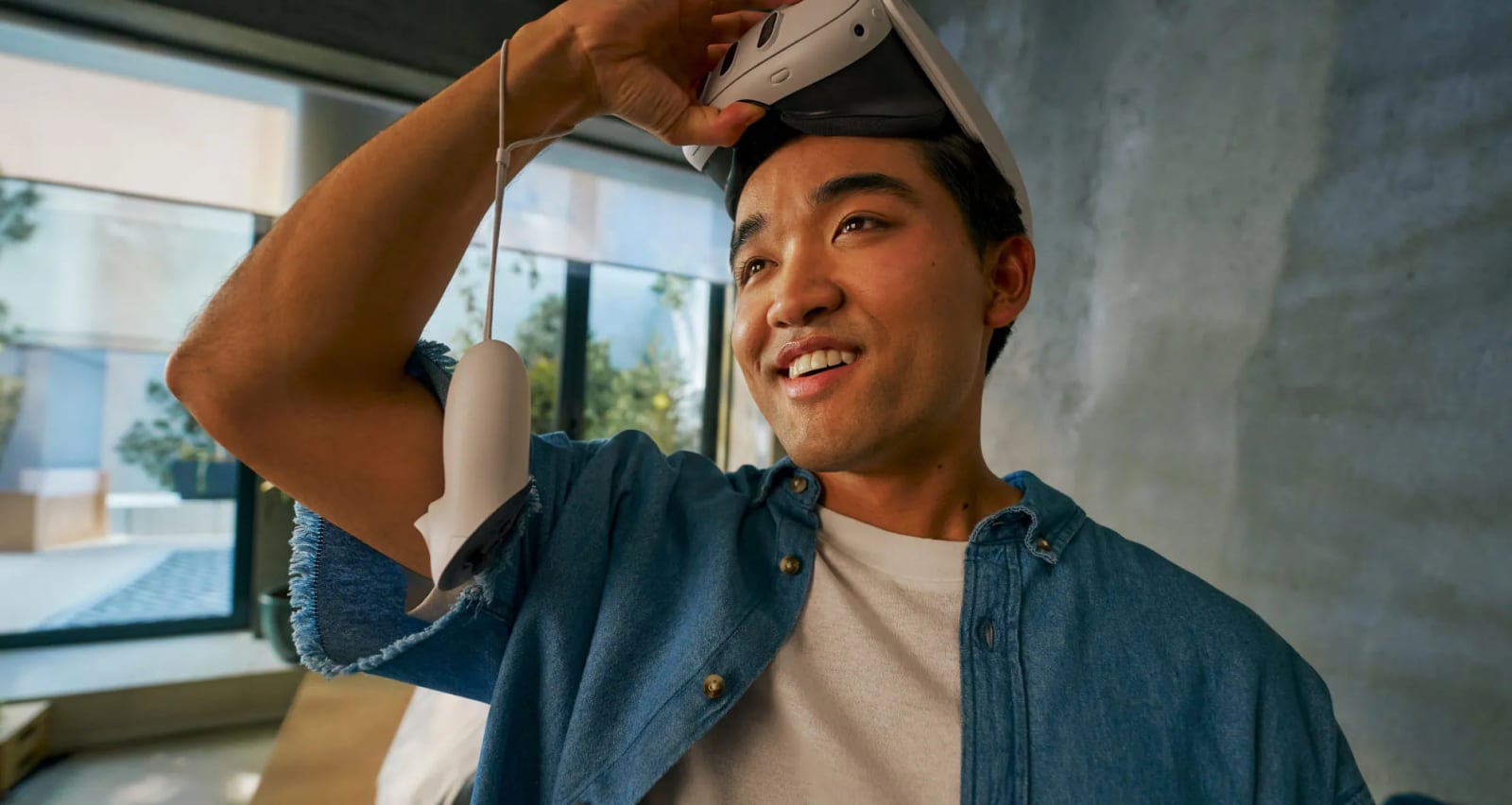 A smiling man lifting a Meta Quest 3 up onto his forehead, a Touch Plus controller hanging from his wrist by its strap