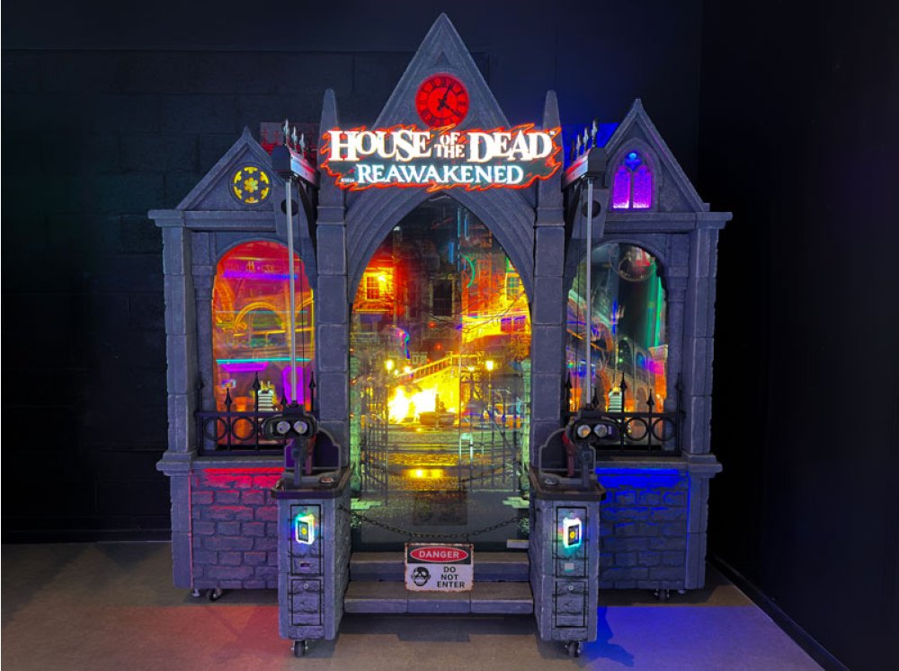 The House of the Dead Reawakened arcade cabinet in a dark room, a grey stone gothic facade with a red clock face above a glowing logo, a large screen in the arched doorway and two arched window screens, with a gun and headset docked on a stone pedestal at each side of the front steps