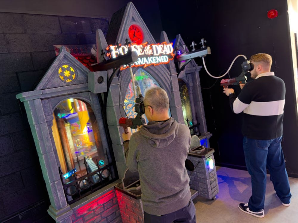 Two men playing House of the Dead Reawakened at the cabinet, each standing in front of the facade holding a red and grey gun with the attached headset raised to his face, cables running down from overhead booms