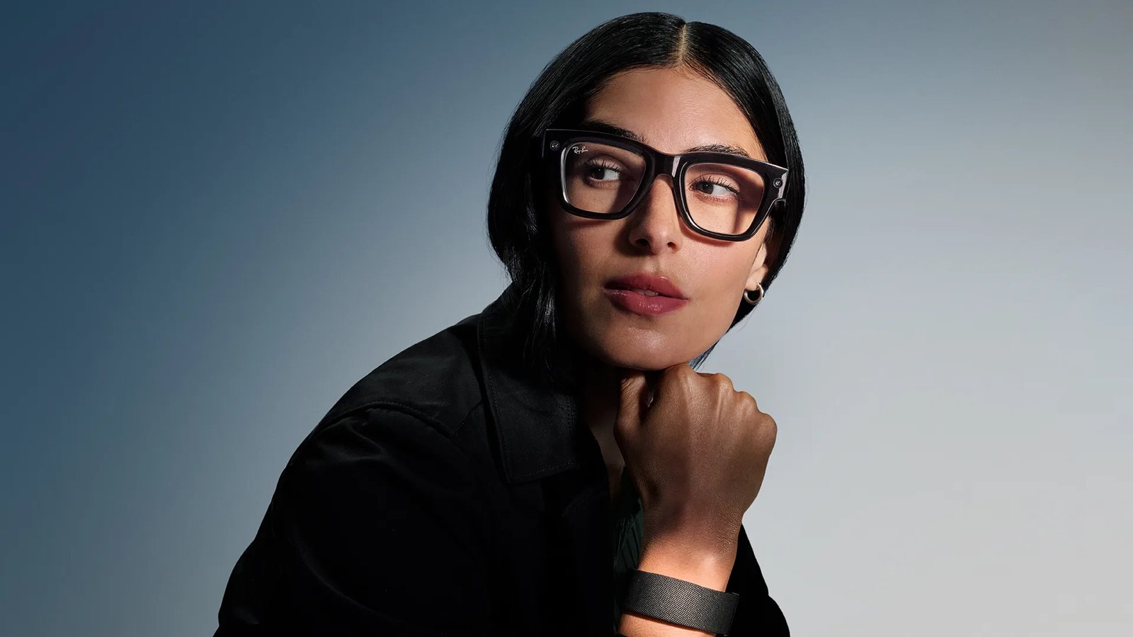 A woman wearing black Meta Ray-Ban Display glasses rests her chin on her fist, with the Meta Neural Band wristband visible on her wrist