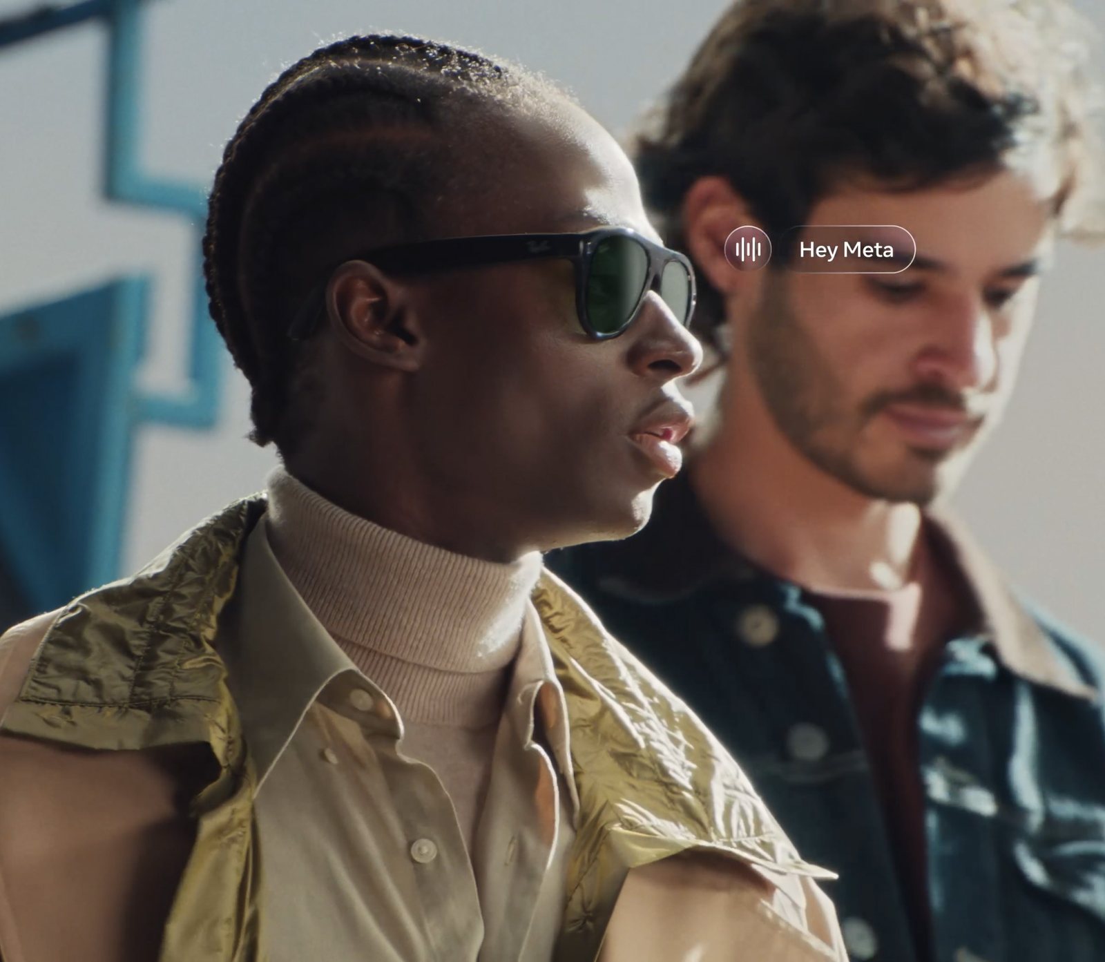 A man wearing dark green-lensed Ray-Ban Meta Gen 3 sunglasses speaks while a Hey Meta voice prompt appears beside him, with a second man behind him