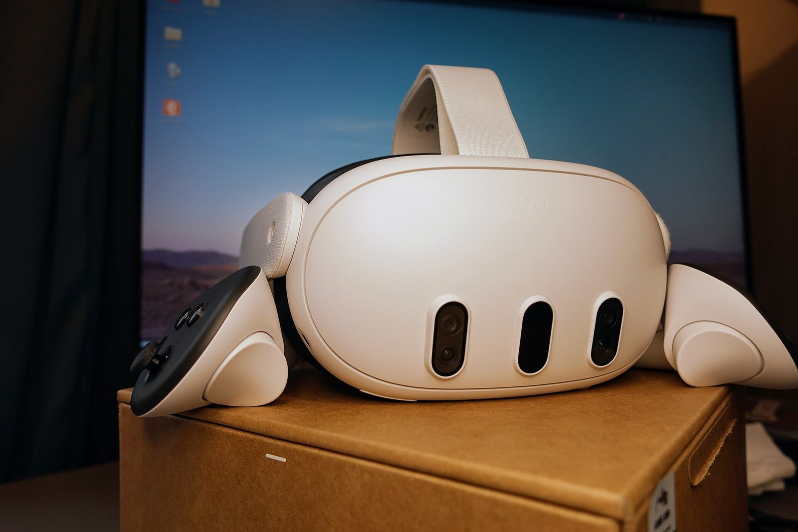 A white Meta Quest 3 headset resting on a cardboard box between its two Touch Plus controllers, with a computer monitor behind it