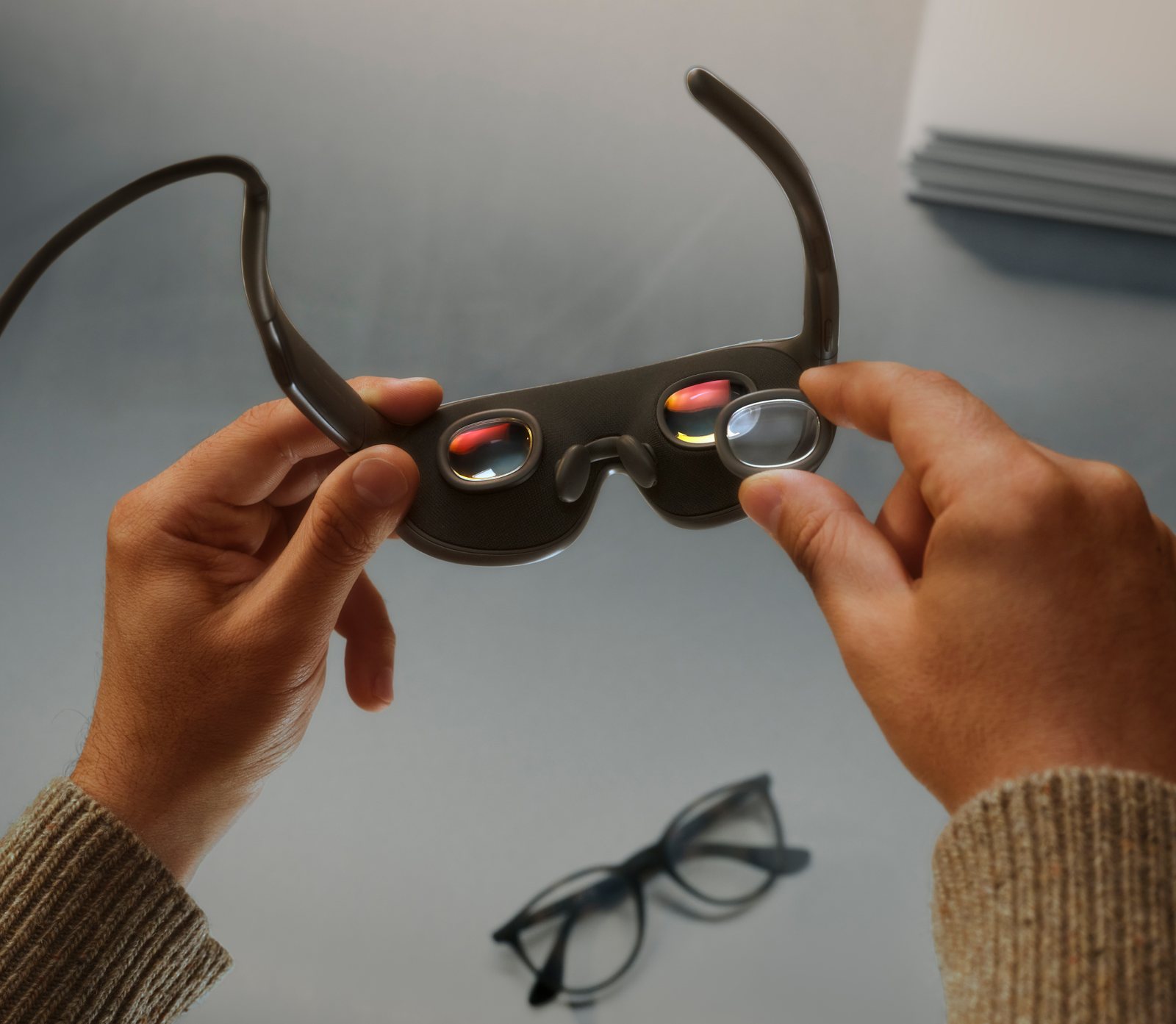 Hands holding Meta VR Glasses from the back, showing two lens openings, nose pads and a clip-in lens insert being fitted, with an ordinary pair of eyeglasses on the table below