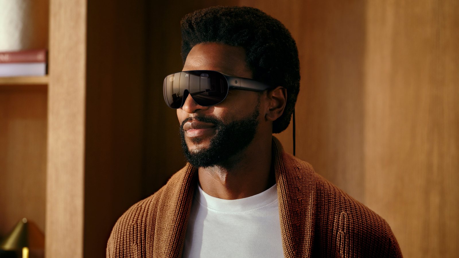 A man in a brown cardigan wearing Meta VR Glasses, a slim dark wraparound visor with temple arms, with a thin cable running down behind his ear