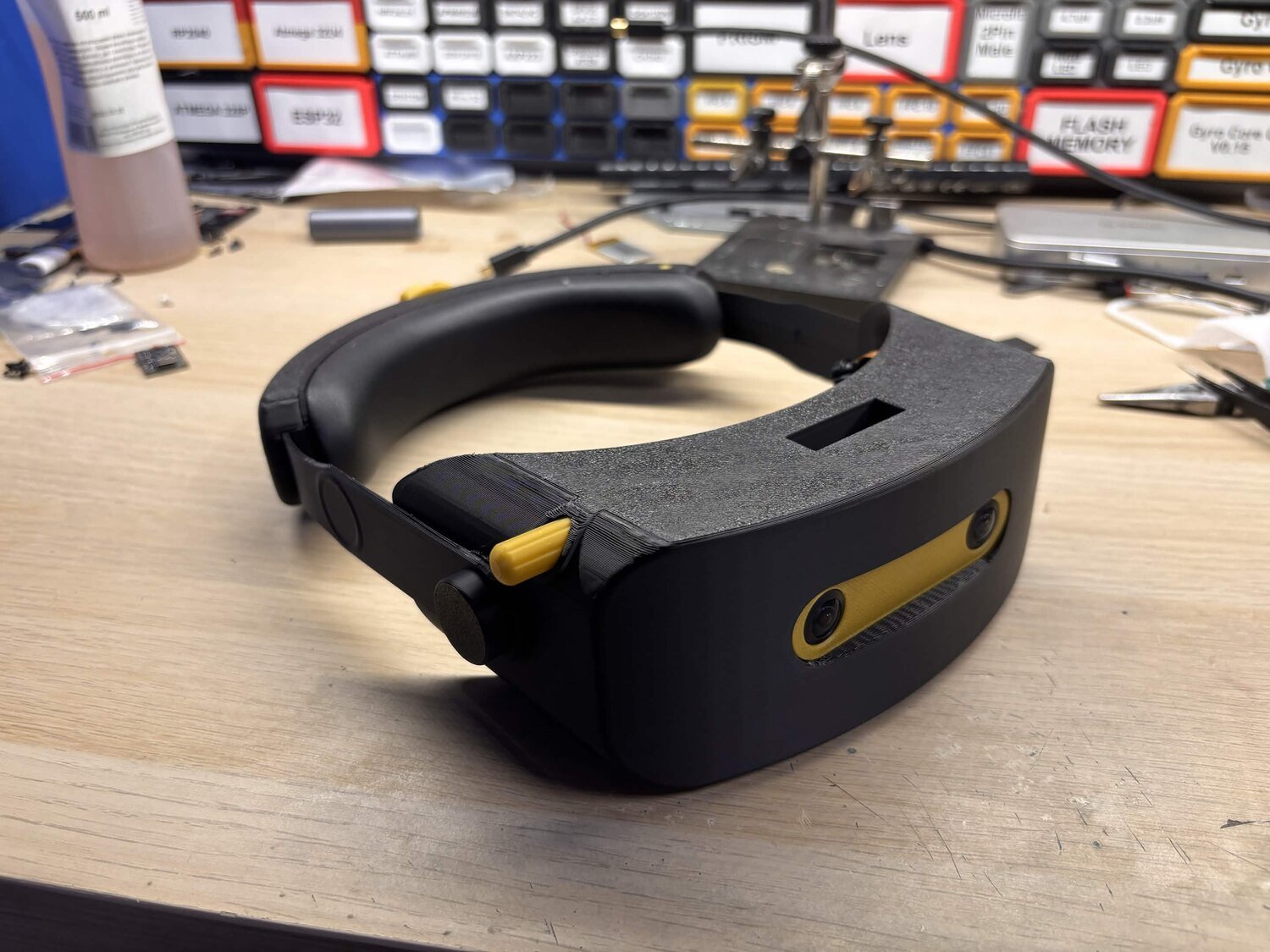 A black 3D-printed Andromeda prototype headset with yellow accents on a workbench, showing a yellow-framed camera strip on the front and a padded halo strap, with component drawers behind it
